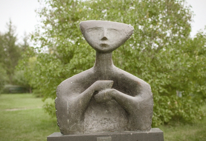 Abstrakt stenskulptur av en människofigur med stiliserade drag, placerad utomhus med träd i bakgrunden.