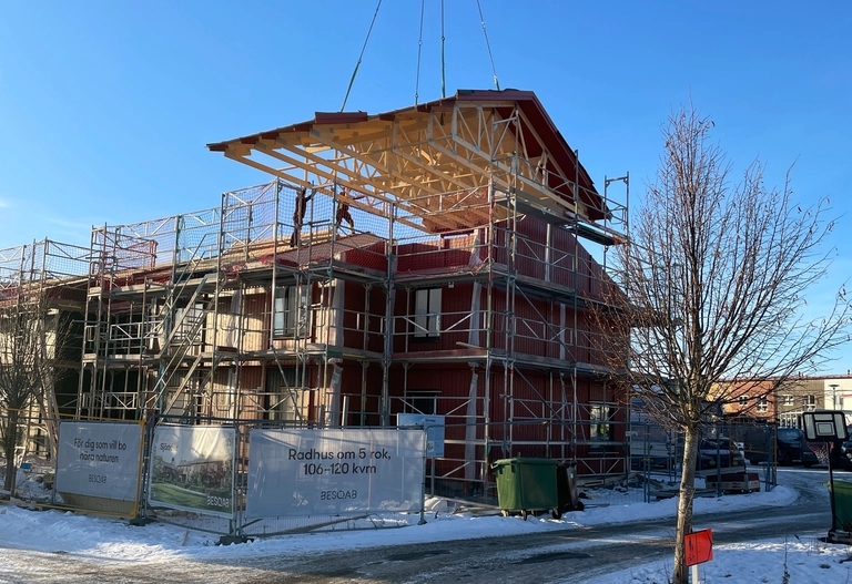 En kran lyfter en taksektion på en delvis uppförd byggnad som omges av byggnadsställningar under en klar dag.