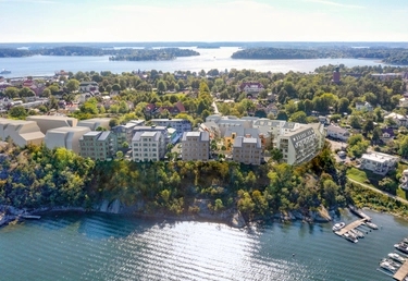 Moderna bostadshus på en lummig kulle vid vattnet, med småbåtar och en stad i bakgrunden under en solig himmel.