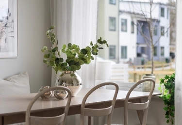 En ljus matplats med ett träbord, stolar och en vas med gröna löv vid ett fönster med utsikt över bostadsområdet..