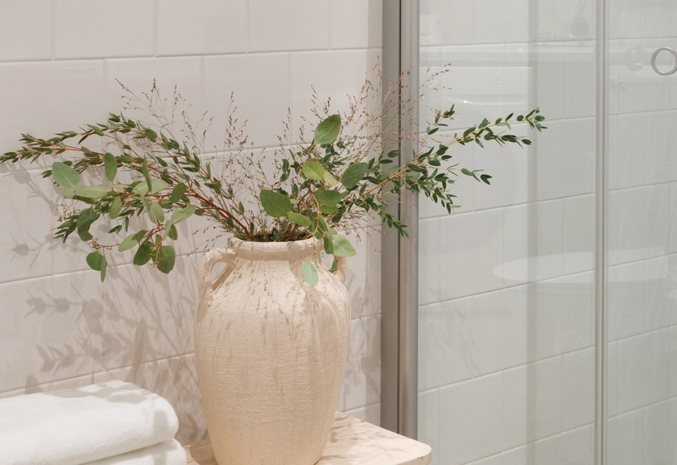 Beige texturerad vas med gröna bladgrenar och torkat gräs, vid vitkaklad vägg och dusch med vita handdukar.