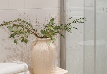 Beige texturerad vas med gröna bladgrenar och torkat gräs, vid vitkaklad vägg och dusch med vita handdukar.