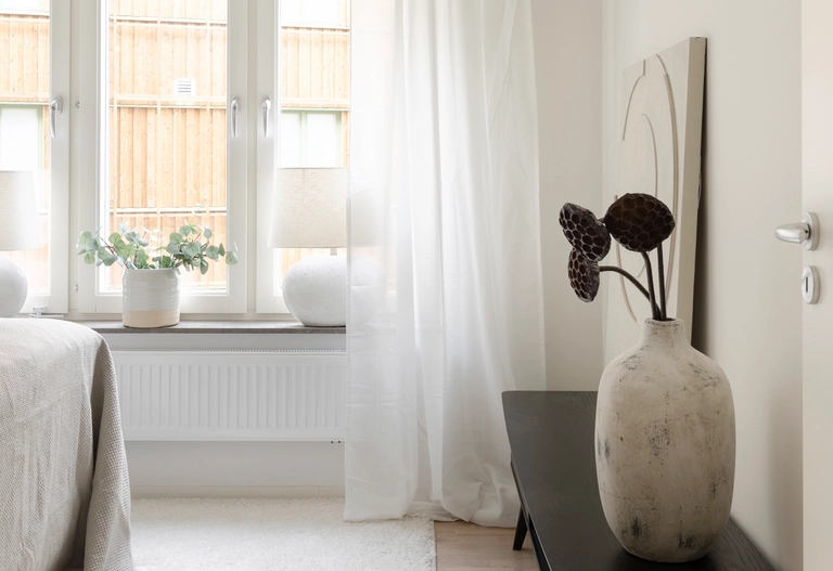 Minimalistiskt sovrum med ljusa väggar, stora fönster, skira gardiner och en keramisk vas med torkade lotusblad på en svart bänk.