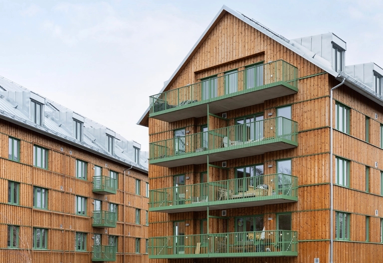 Flerfamiljshus med träpanel på utsidan och gröna metallbalkonger under en molnig himmel.