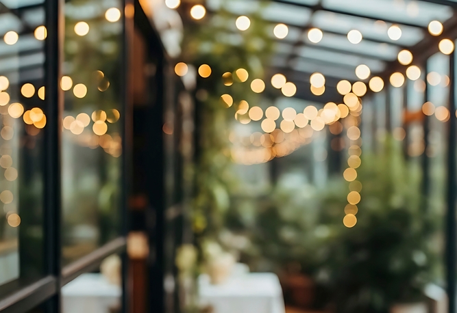 Ljusslingor som hänger i ett rum med glasväggar, omgivna av grönska och suddiga bord i bakgrunden.