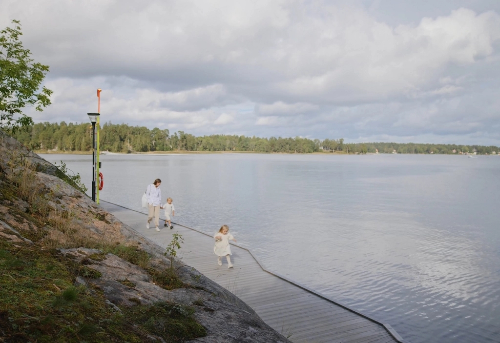 Tre personer går längs en träbrygga vid ett lugnt vatten, med träd och klippor i bakgrunden under en molnig himmel.