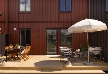 Rött trähus med trädäck. Till vänster matgrupp, till höger loungemöbler under vitt parasoll.
