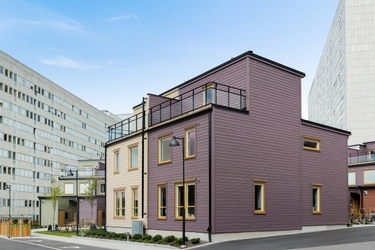 Modernt radhus i mörklila och beige med takterrass, omgivet av höghus i en stadsmiljö under blå himmel.