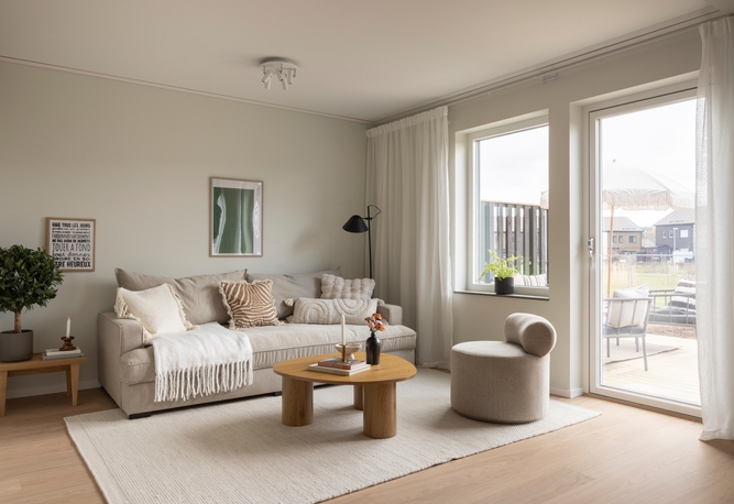 Ljust och modernt vardagsrum med beige L-soffa, lågt soffbord, ottoman, matta och stora glasdörrar mot en uteplats.