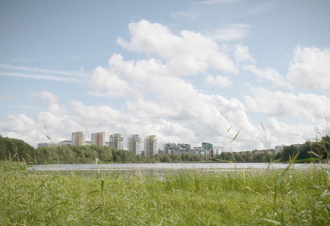 Ett gräsbevuxet fält vid en sjö med en stadssilhuett och moln i bakgrunden.