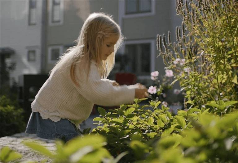 Ung flicka med långt blont hår plockar eller undersöker växter i en trädgård, med byggnader i bakgrunden.