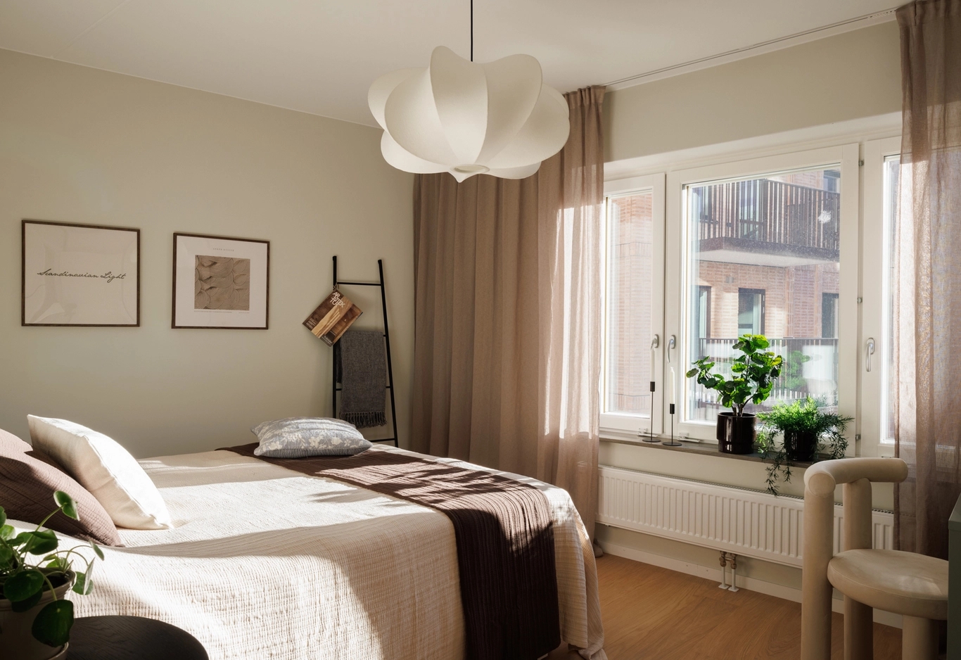 Sovrum med beige väggar, säng med vita och bruna täcken, två tavelramar, stort fönster med bruna gardiner, växter och en vit organisk taklampa.