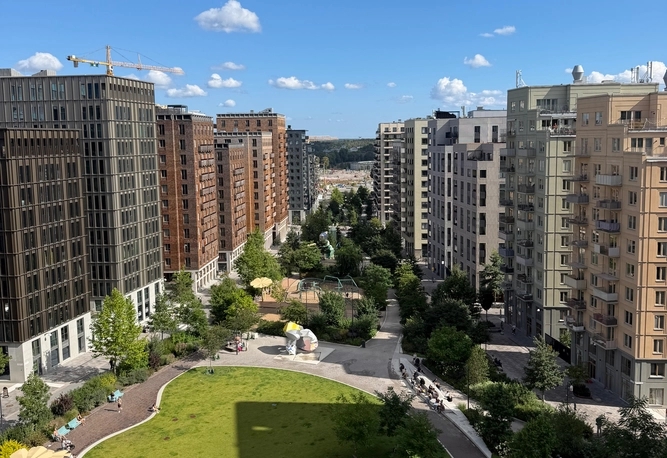 Vy över Norra stationsparken. Flera höga, moderna lägenhetshus runt en grön park med gångvägar och träd under en blå himmel.