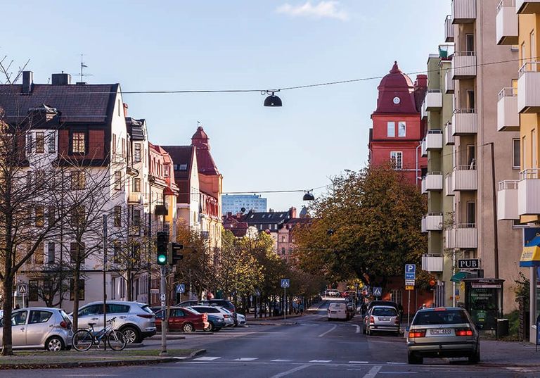 En stadsgata med parkerade bilar, träd och byggnader på båda sidor under en klar himmel i dagsljus.
