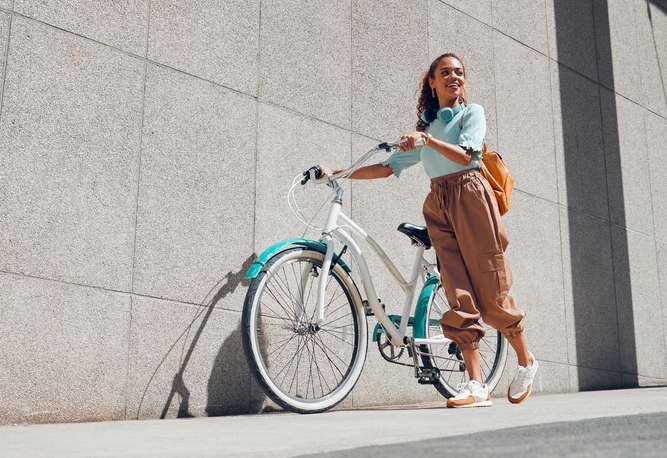Leende kvinna i ljusblå topp och beige byxor står med vit cykel, blå skärmar, framför grå vägg. Gul ryggsäck.
