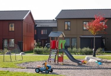 Lekplats med rutschkana och sandlåda på gården, framför radhus i rött, svart och olivgrönt trä