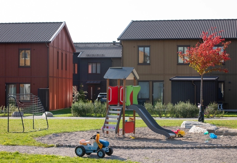 Lekplats med rutschkana och sandlåda på gården, framför radhus i rött, svart och olivgrönt trä