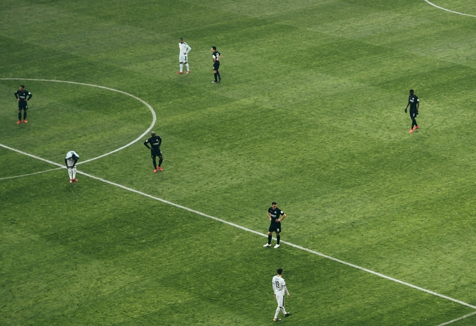 Grön fotbollsplan med vita linjer. Flera spelare i svarta och vita uniformer står på planen. En fotboll ligger vid en spelares fötter.