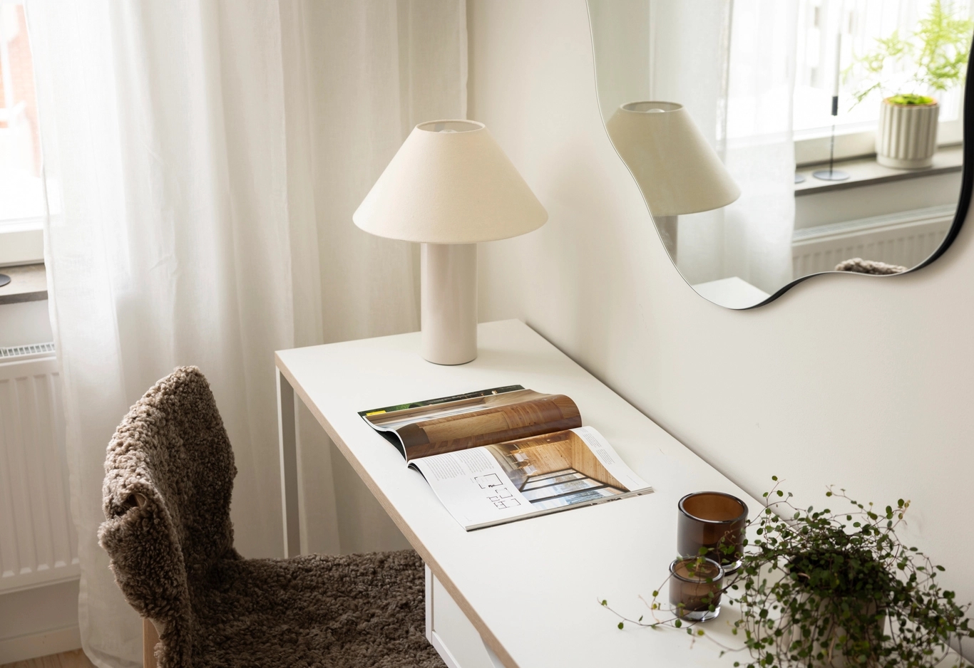 Ett vitt skrivbord med en uppslagen tidning, en lampa, en liten växt och två stearinljus står vid ett fönster med en vågig spegel ovanför.