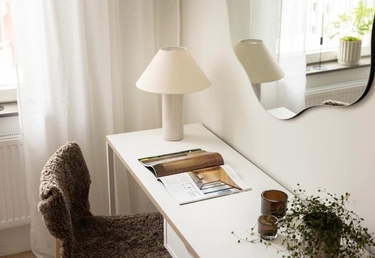 Ett vitt skrivbord med en uppslagen tidning, en lampa, en liten växt och två stearinljus står vid ett fönster med en vågig spegel ovanför.