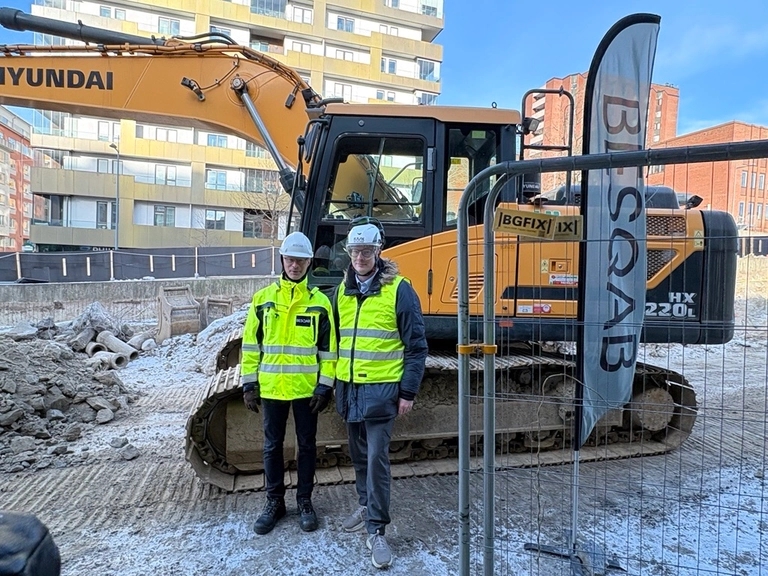 Två personer i varseljackor och skyddshjälmar står framför en Hyundai-grävmaskin på en byggarbetsplats.