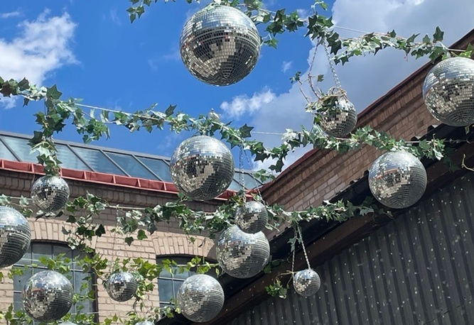 Glänsande discokulor och gröna lövverksgirlanger över en innergård mot blå himmel och byggnader.