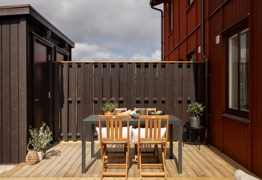 Uteplats i trä med mörkt trästaket, rött hus och förråd. Grått bord med trästolar och krukväxter under molnig himmel.