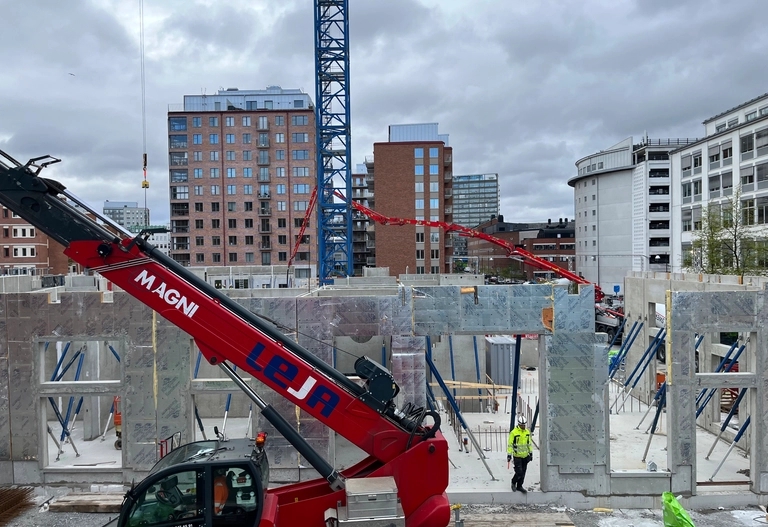Byggarbetsplats med kranar, betongväggar och arbetare; flervåningshus och molnig himmel i bakgrunden.