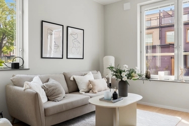 Ljust vardagsrum med beige soffa, två tavlor, ljust soffbord med blommor och stora fönster mot grönska och byggnader.