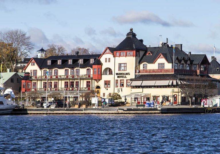 Historiskt hotell vid vattnet med röda tak och uteservering, beläget bredvid en flod under en delvis molnig himmel.