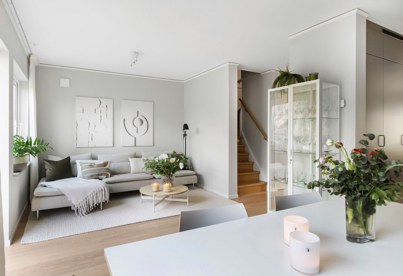Ett vardagsrum med en grå soffa, vit konst på väggarna, ett vitrinskåp, en trätrappa och ett matbord med ljus och blommor i förgrunden.