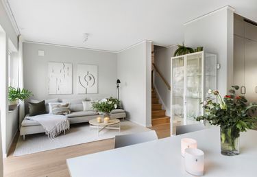 Ett vardagsrum med en grå soffa, vit konst på väggarna, ett vitrinskåp, en trätrappa och ett matbord med ljus och blommor i förgrunden.