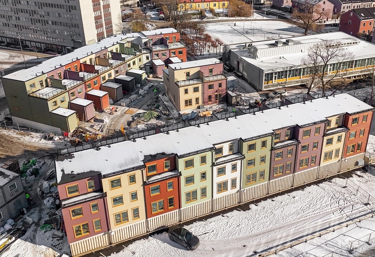 Flygfoto över färgglada radhus och närliggande byggnader med snö som täcker marken i ett stadsområde.