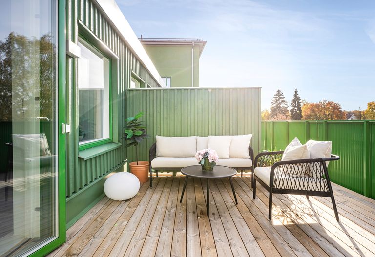 Takterrass med trädäck mot grön väggfasad. Vita kuddar på en soffa, svart runt bord, flätad stol och krukväxt i solljus.
