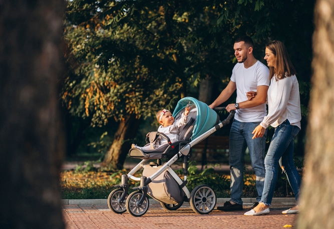 En man och en kvinna går tillsammans utomhus och skjutsar ett barn i en barnvagn längs en gångväg med träd i närheten.