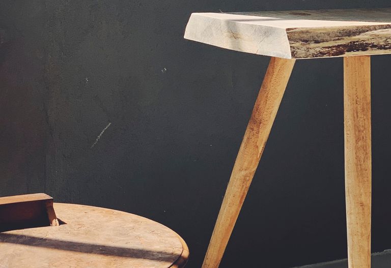 Närbild av ett träbord med vinklade ben bredvid en rund träpall mot en mörk vägg.