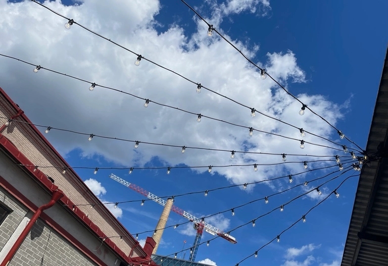 Ljusslingor spända över en blå himmel med vita moln mellan byggnader. En rödvit byggkran syns i bakgrunden.
