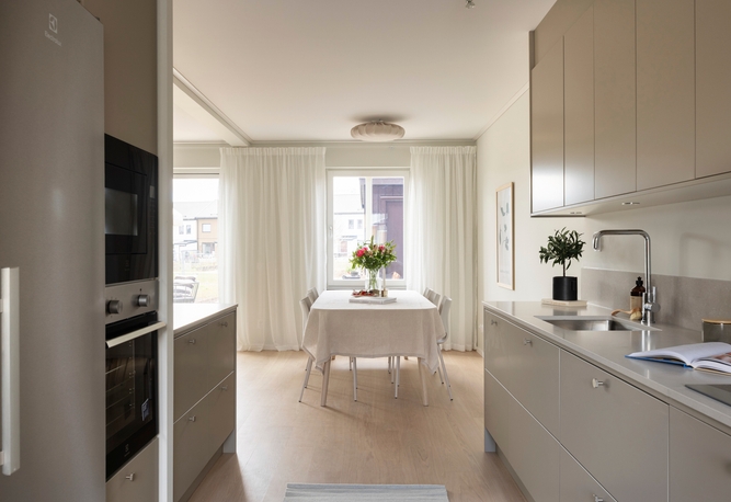 Ljust kök och matplats i modern stil. Beige skåp, vitklätt matbord med blommor vid fönster med vita gardiner. Ljust trägolv.