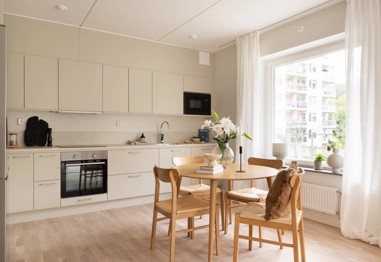 En terrass med trämöbler, inklusive ett runt bord, korgstolar och en mysig sittgrupp med en soffa, kuddar och en matta. Växter och grönska pryder utrymmet.