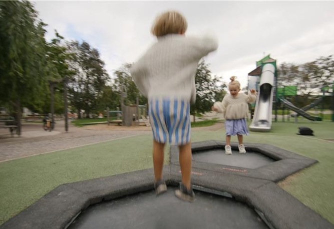 Två barn i randiga shorts leker på sexkantiga trampoliner på en utomhuslekplats med en rutschkana i bakgrunden.