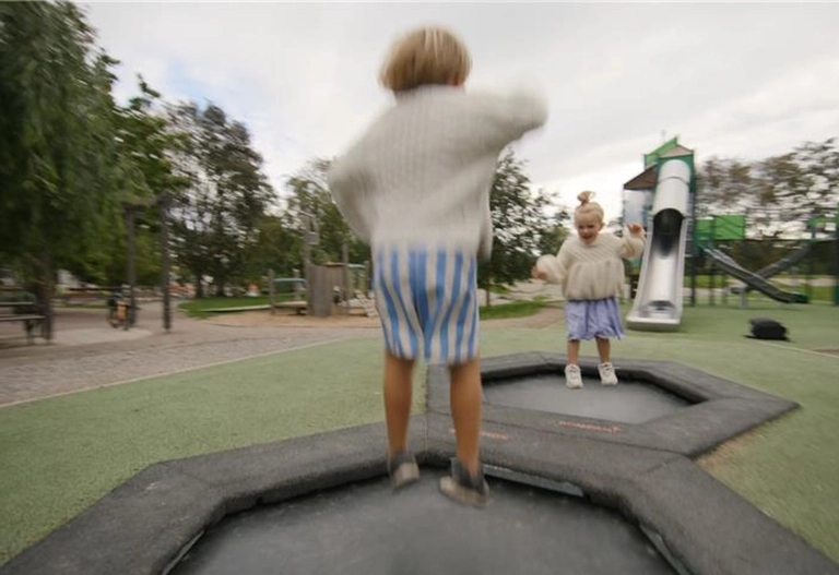Två barn i randiga shorts leker på sexkantiga trampoliner på en utomhuslekplats med en rutschkana i bakgrunden.