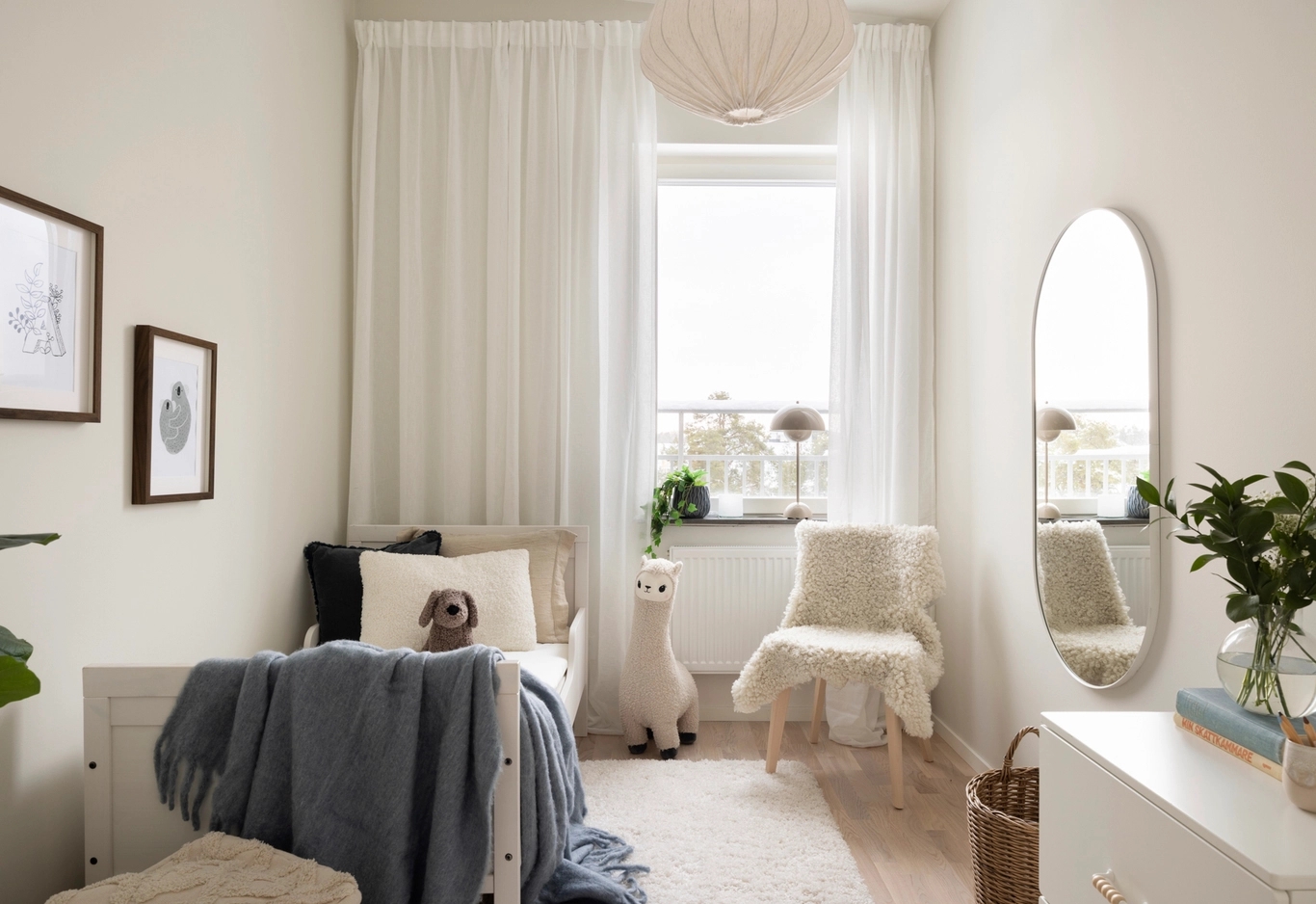 Minimalistiskt sovrum med neutral inredning, en enkelsäng, plyschleksaker, en fåtölj, oval spegel och ett fönster med skira gardiner.
