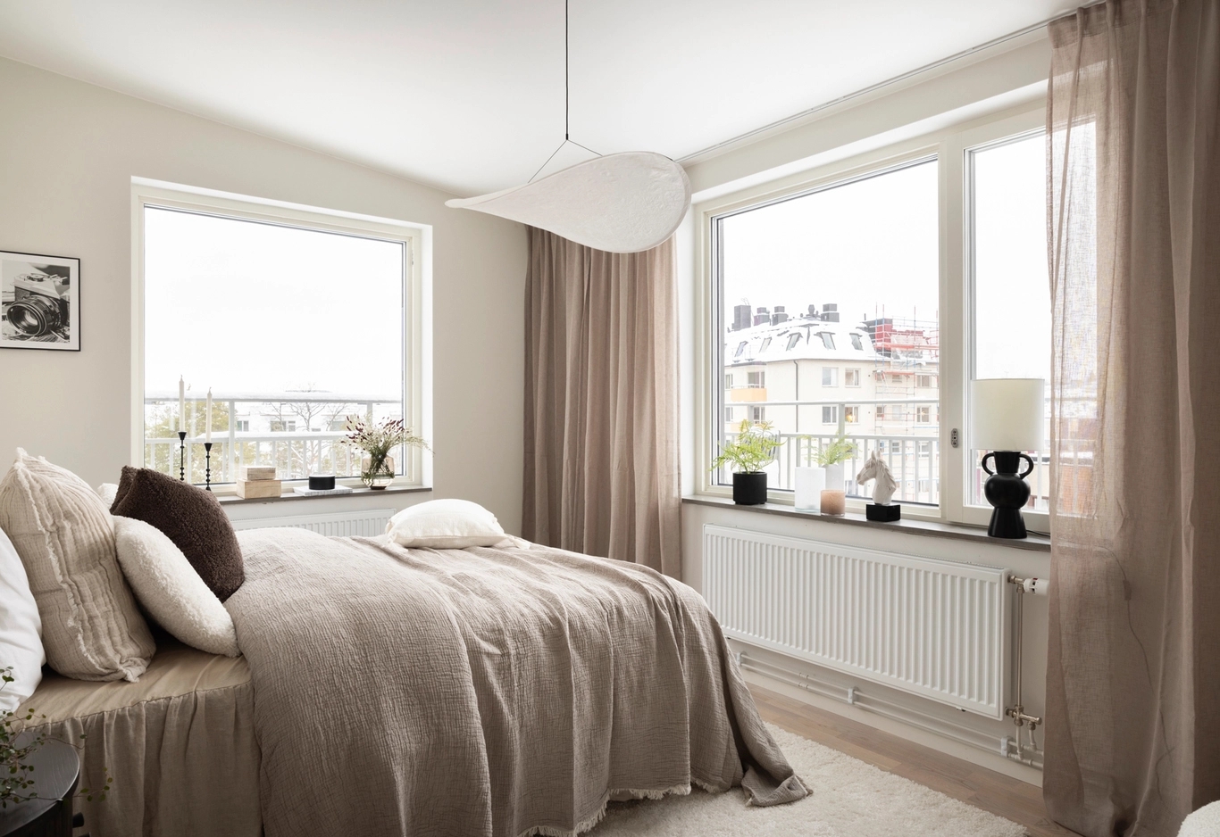 Ljust sovrum med beige sängkläder, stora fönster, skira gardiner och krukväxter på fönsterbrädan.