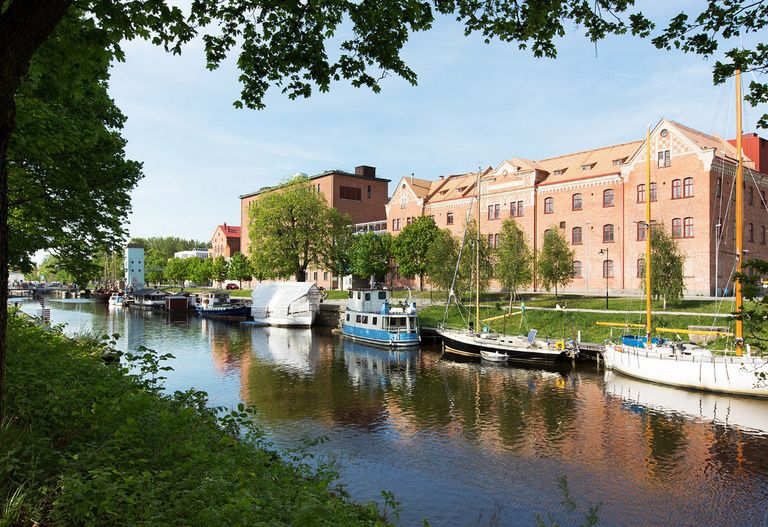 Historiska röda tegelbyggnader längs en å med förtöjda båtar och grönskande träd.