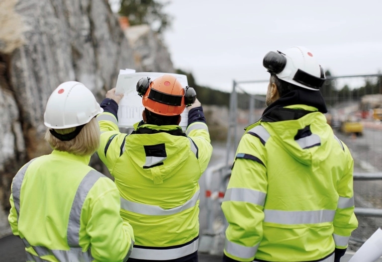 Tre människor står i varselkläder på en byggarbetsplats och tittar på en ritning som en av dem håller upp i luften.