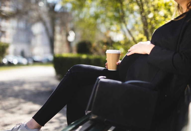 Gravid kvinna som sitter på en bänk i en park i stadsmiljö och dricker en kopp kaffe