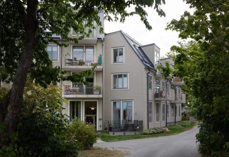 Moderna ljusa lägenhetshus med balkonger bland träd och grönska, med en gångväg i förgrunden.