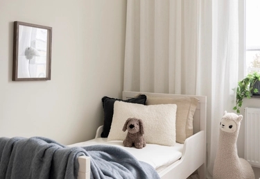 En liten säng med kuddar, en uppstoppad hundleksak och en plyschlama i ett mjukt upplyst rum i neutrala färger.