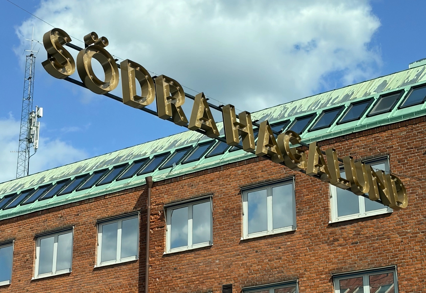Stor guldskylt med texten SÖDRA HAGALUND hänger över en röd tegelbyggnad med vita fönster, under blå himmel.