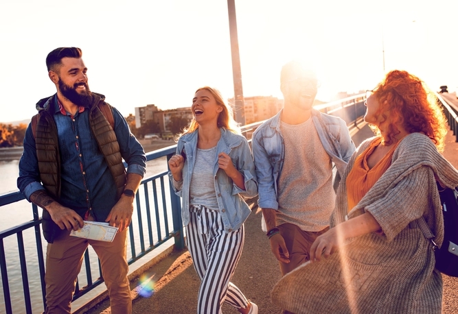 Fyra personer går och pratar tillsammans på en bro i solnedgången, klädda i fritidskläder, med solljus i bakgrunden.